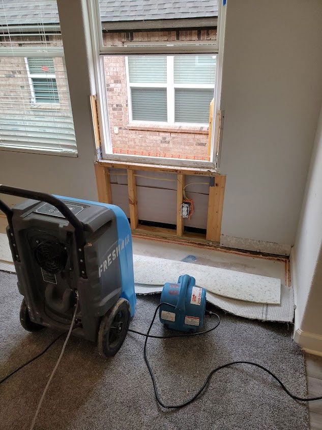 A room under restoration with a large blue industrial dehumidifier and a small fan near a wall with removed drywall.