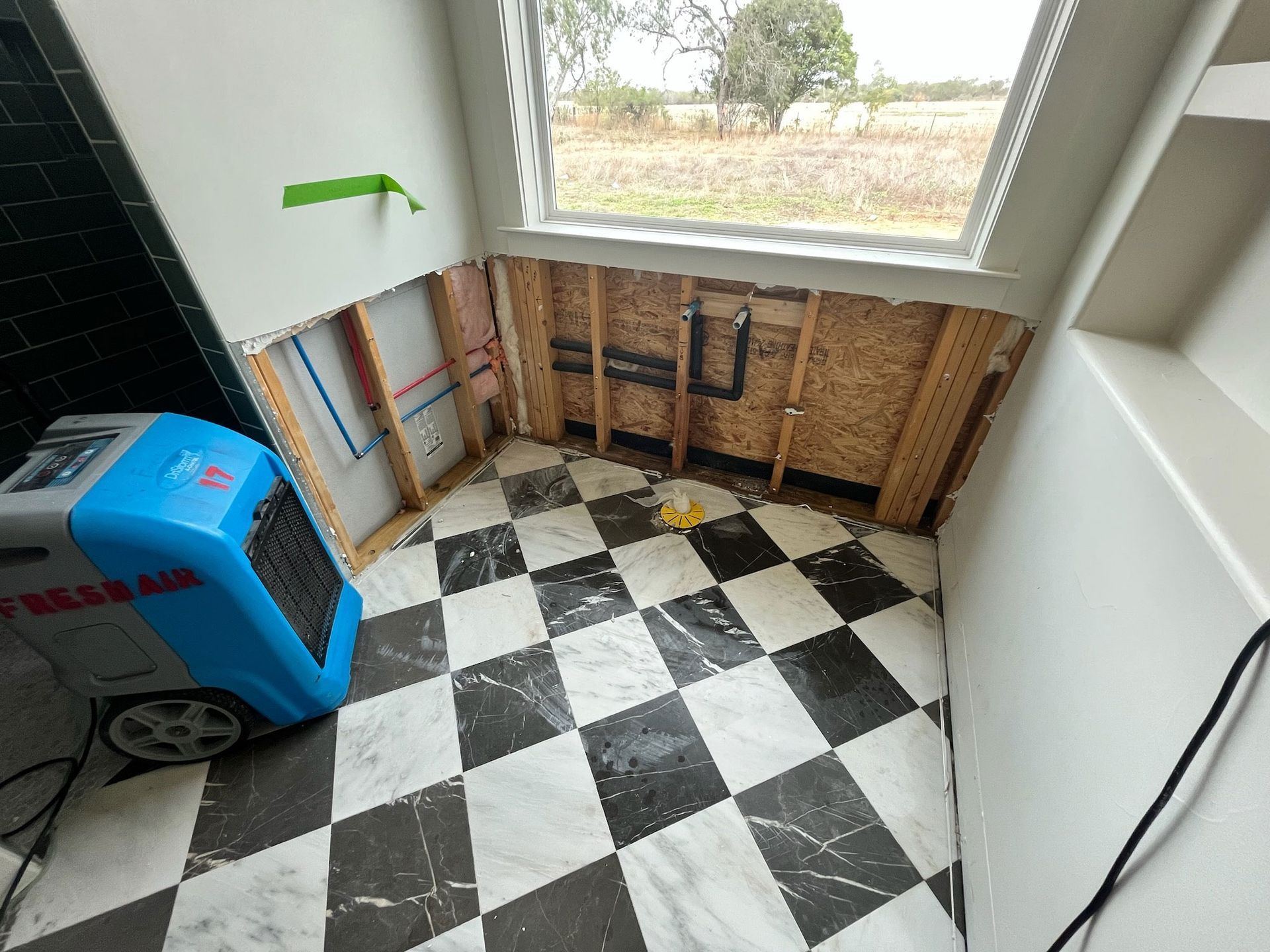 Dehumidifier in a room with walls partially removed to expose wooden studs and plumbing, featuring black-and-white tile.