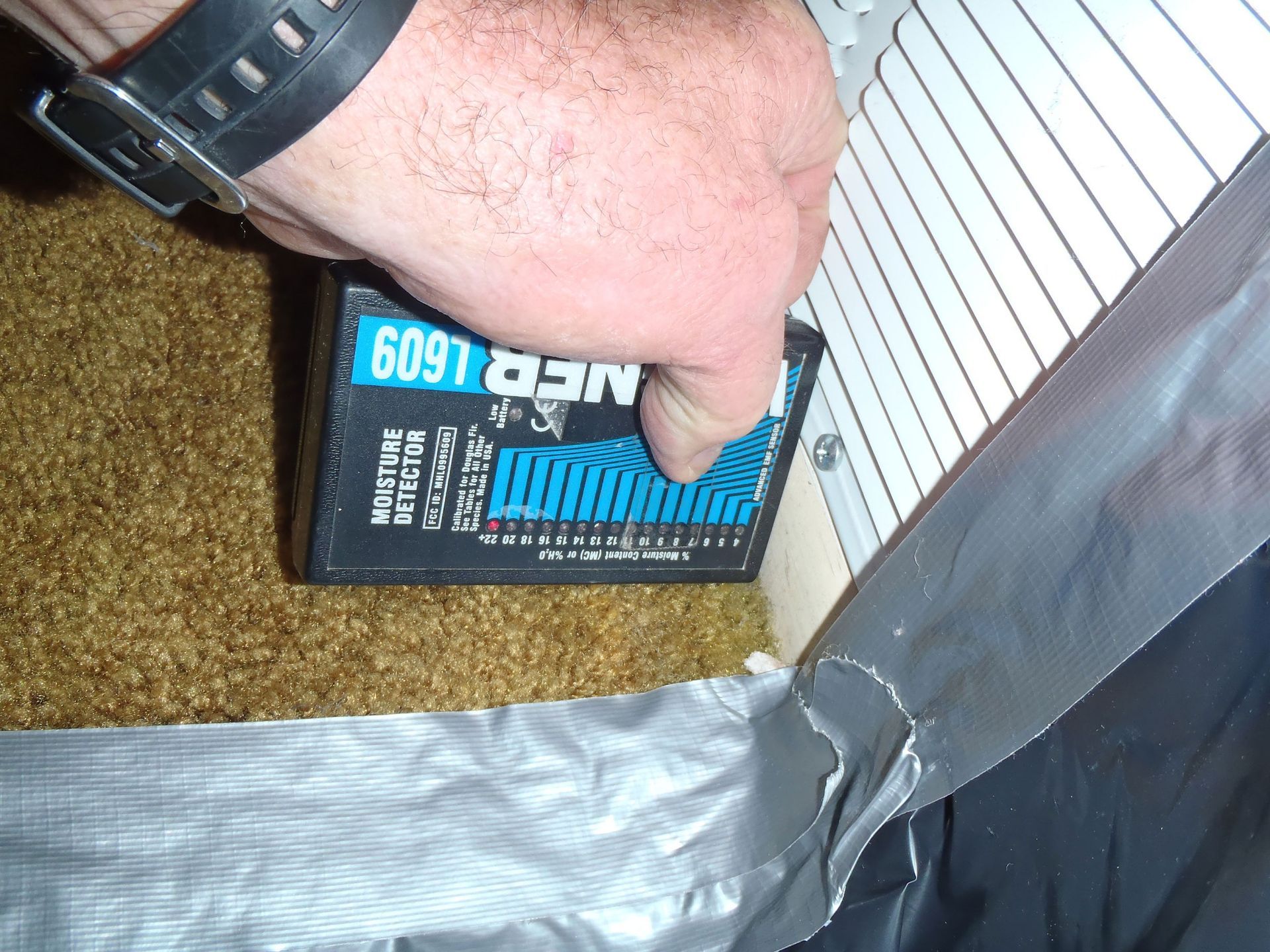 A hand uses a Wagner L609 moisture meter on a carpeted floor next to a white louvered vent with silver duct tape.