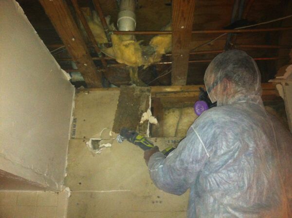 A person in a protective suit and mask uses a tool to cut into a residential ceiling with exposed wooden joists.