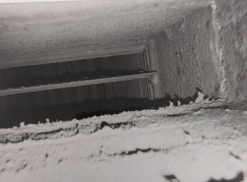 A close-up view inside a dusty, gray ventilation duct showing buildup of debris and a metal damper blade.