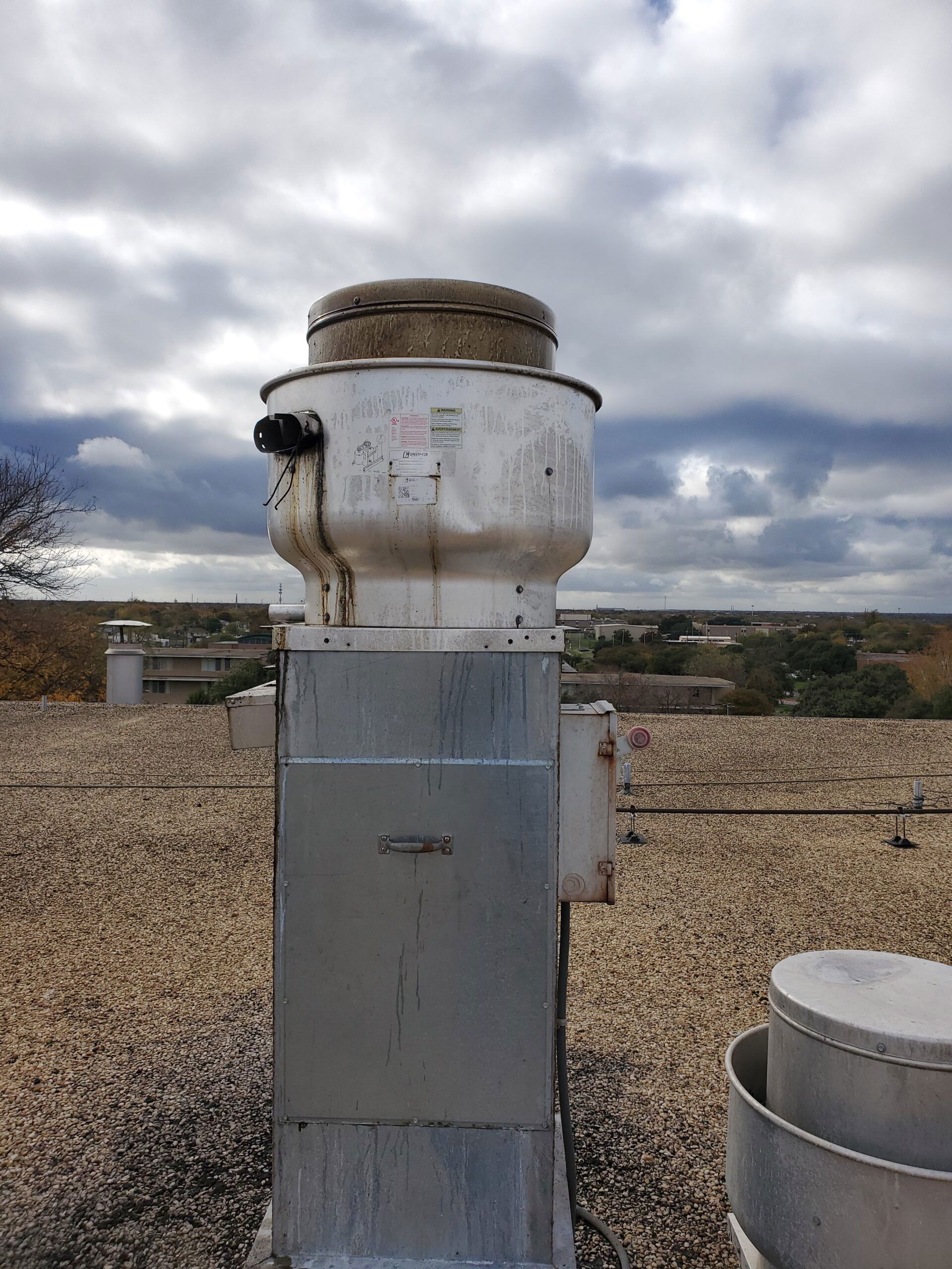 A weathered, metallic rooftop ventilation exhaust fan standing on a gravel-covered flat roof under a cloudy sky.