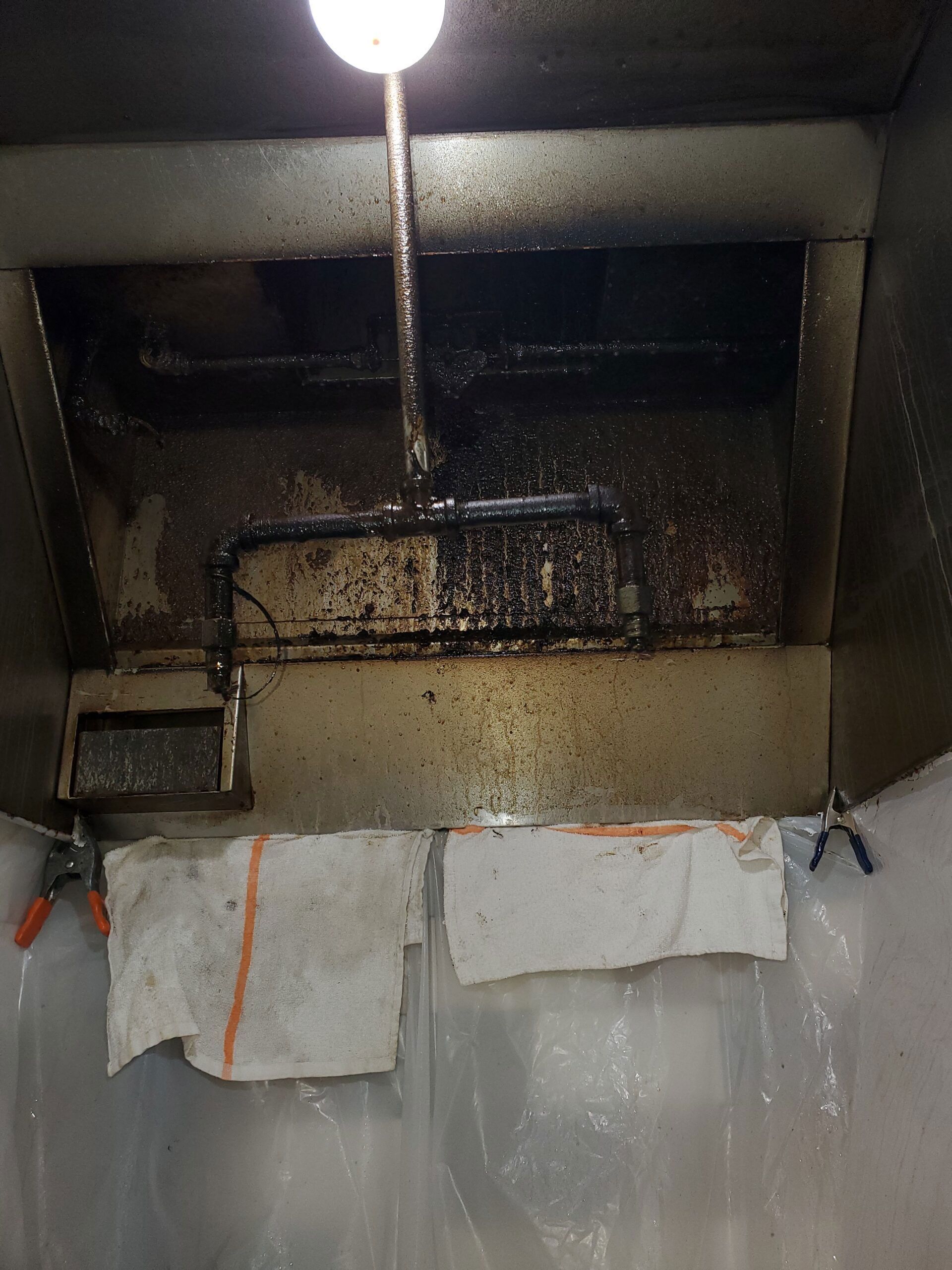 A greasy kitchen exhaust hood vent being deep-cleaned with protective plastic sheeting and white cloths attached.