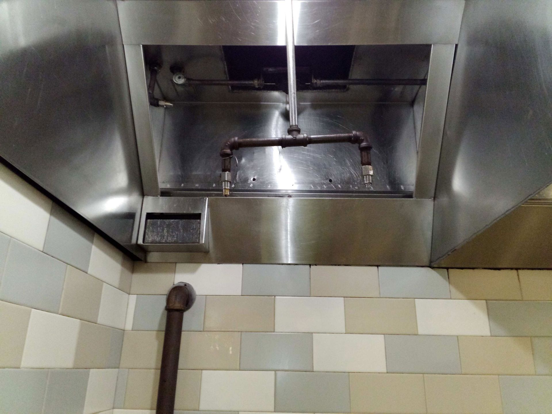 A commercial stainless steel kitchen exhaust hood with an internal pipe system against a tiled wall.