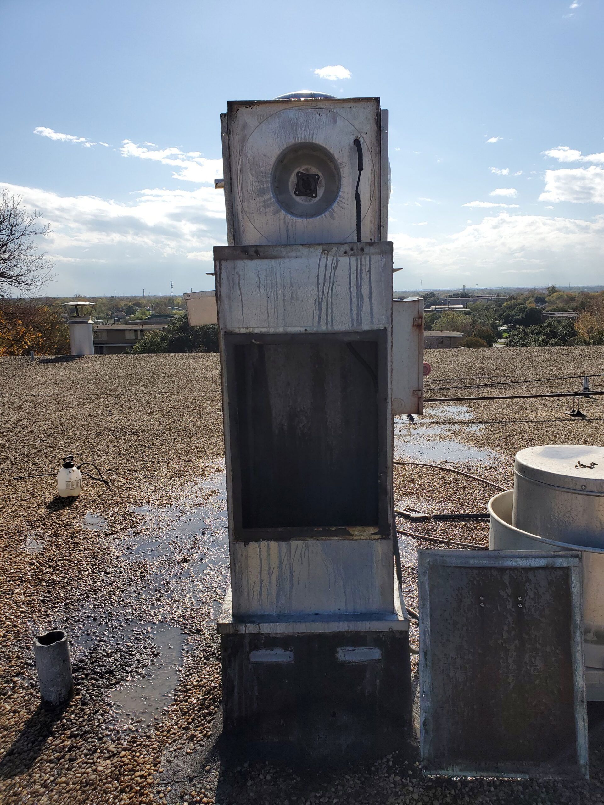 A weathered, boxy industrial air handling unit stands on a gravel rooftop under a bright, partly cloudy sky.