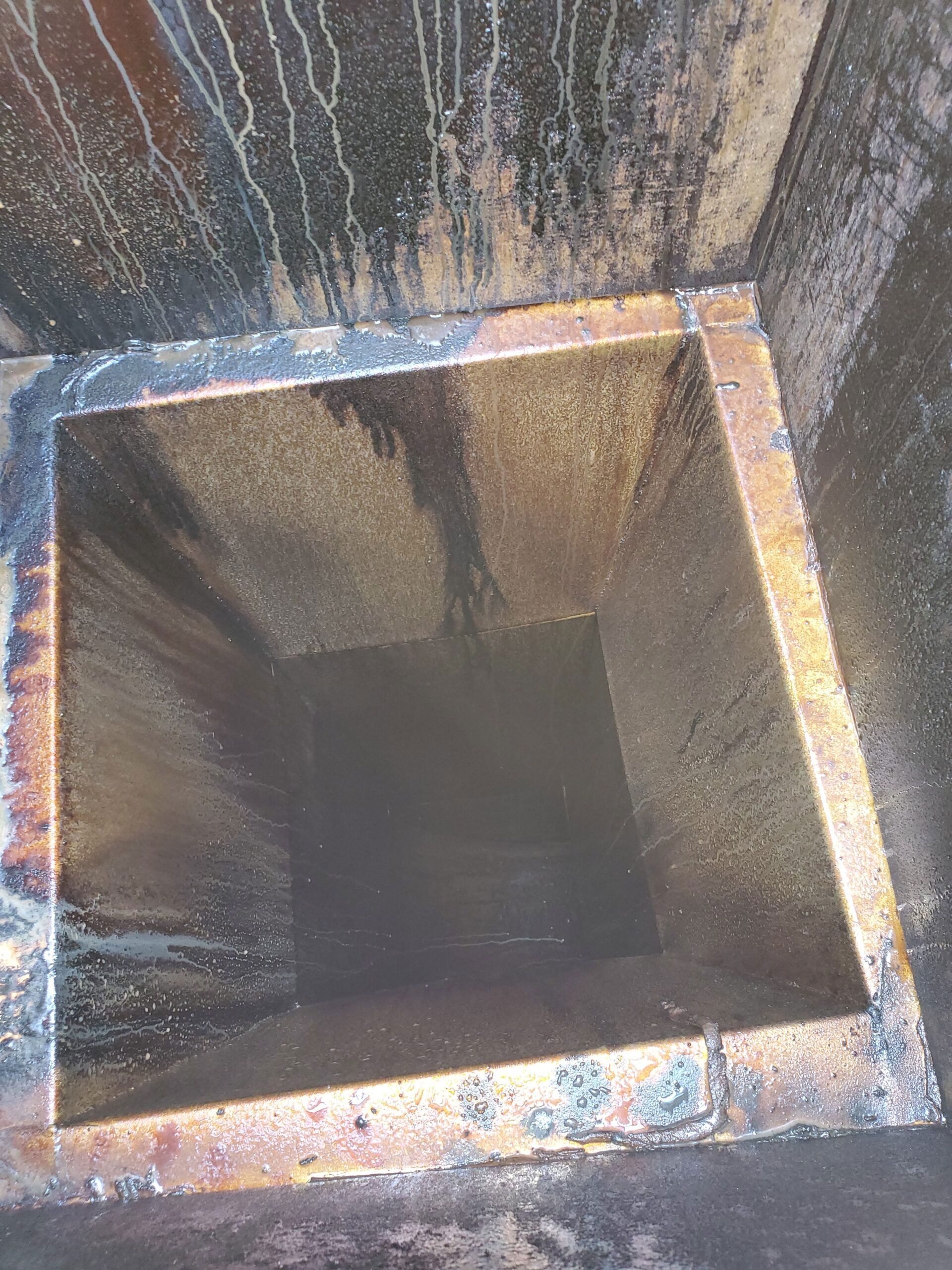 A square metal shaft leading into darkness, with rusted, stained walls and streaky residue from oil or water.