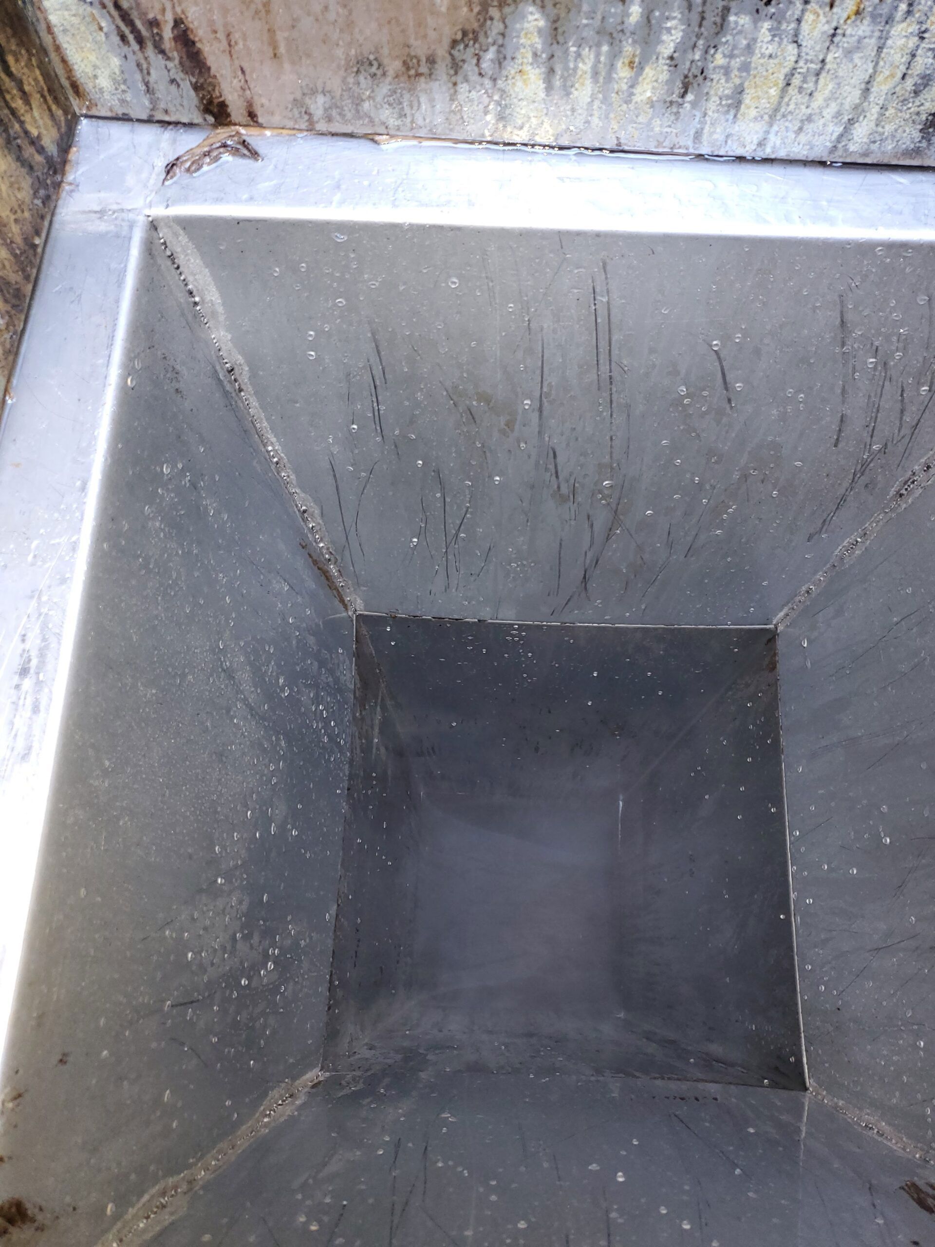 Looking down into a deep, empty square metal drainage shaft with dark, murky water at the bottom.