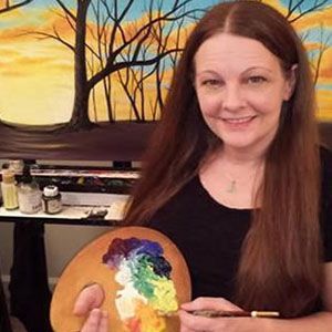 A woman is holding a palette of paint and a brush in front of a painting.