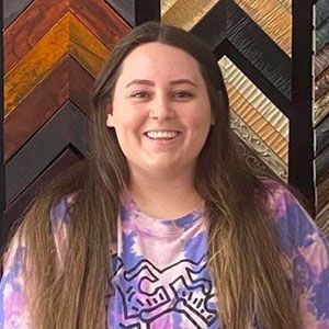 A woman in a purple tie dye shirt is smiling in front of a wall of picture frames.