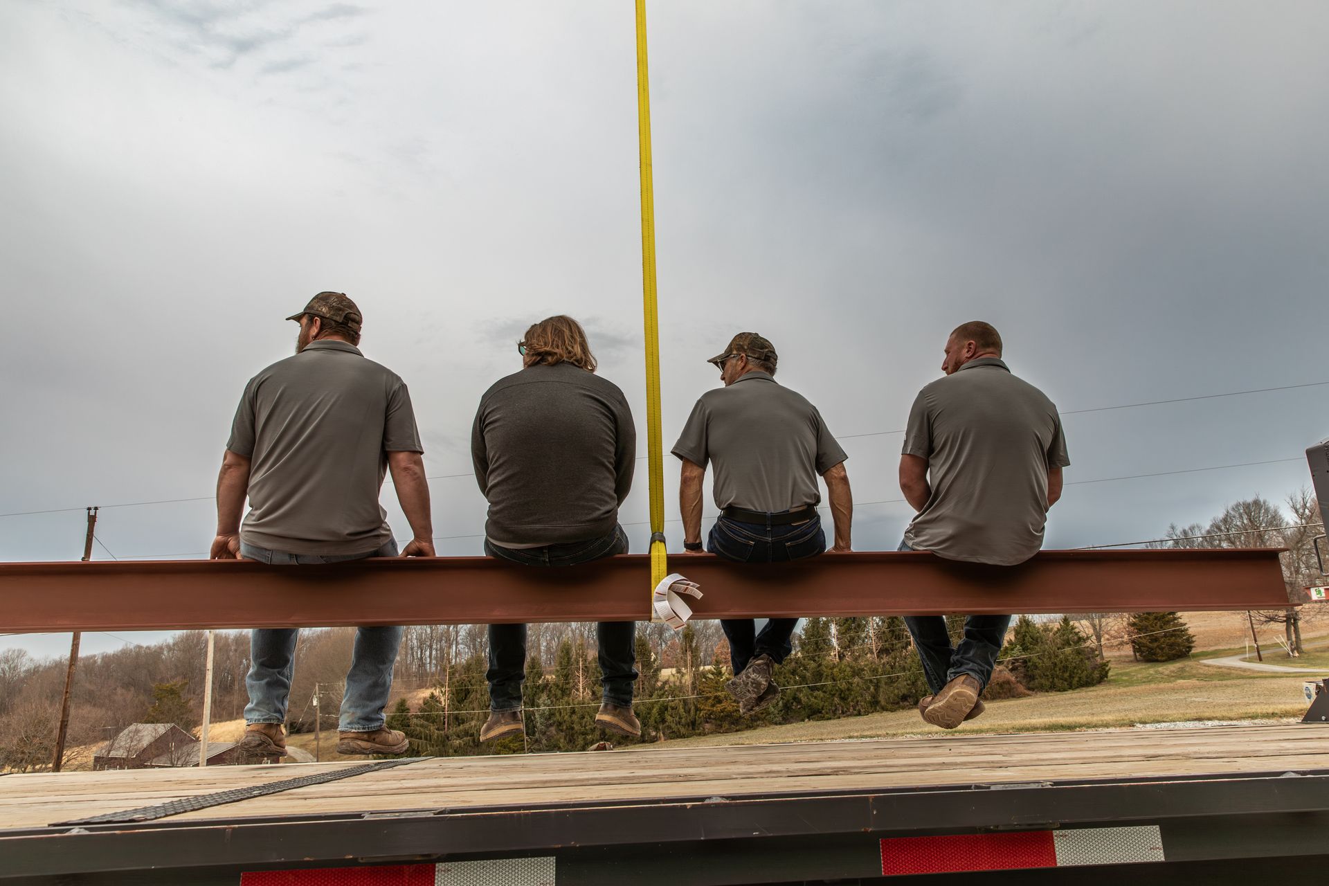 Four people wearing gray shirts sit on a suspended steel beam against a cloudy sky above a flatbed trailer.