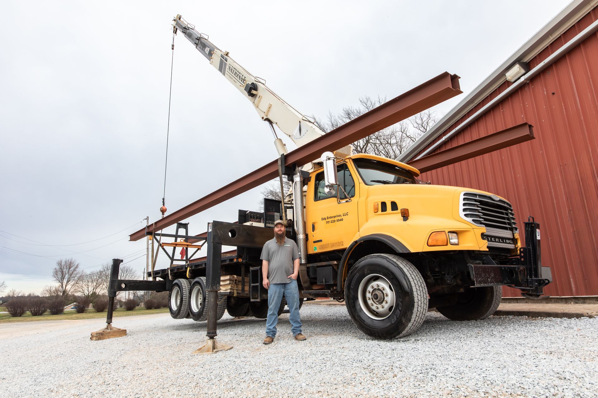 A person stands in front of a yellow boom truck crane carrying a large steel I-beam next to a red building on gravel.