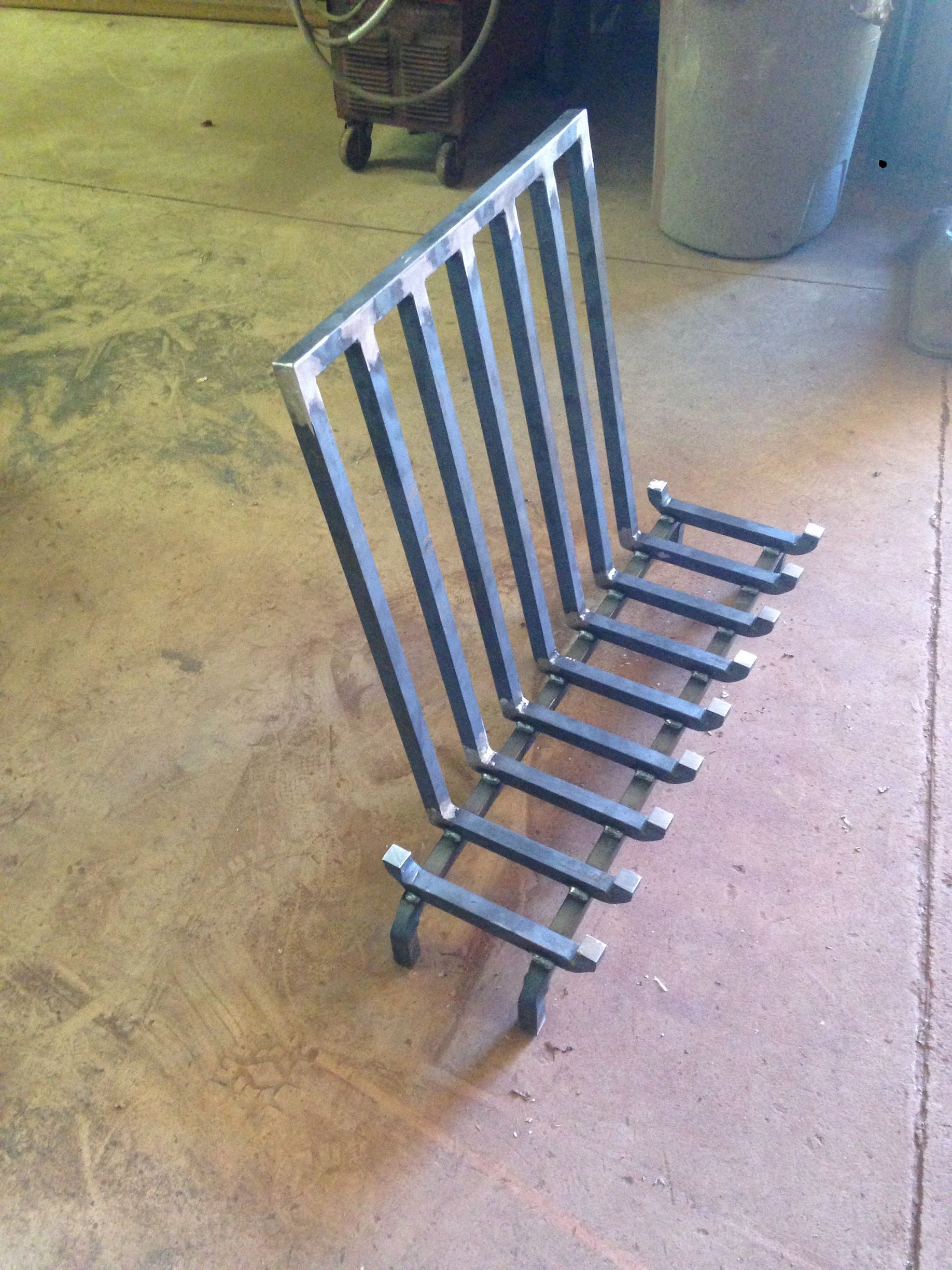 A newly fabricated metal fireplace grate with a rectangular back and a horizontal base with short legs on a concrete floor.