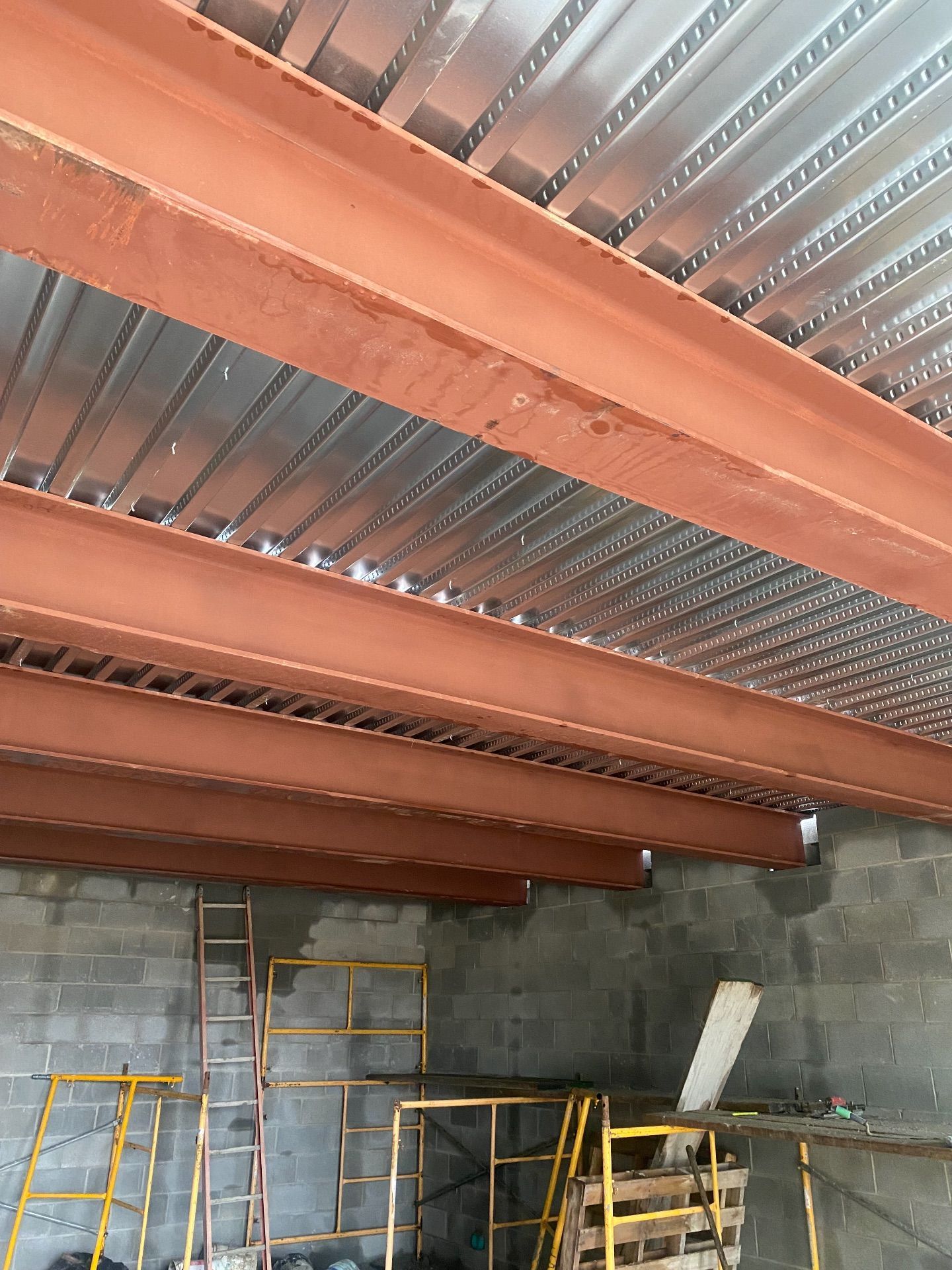 Steel beams support a corrugated metal floor deck above a workspace with scaffolding and cinder block walls.