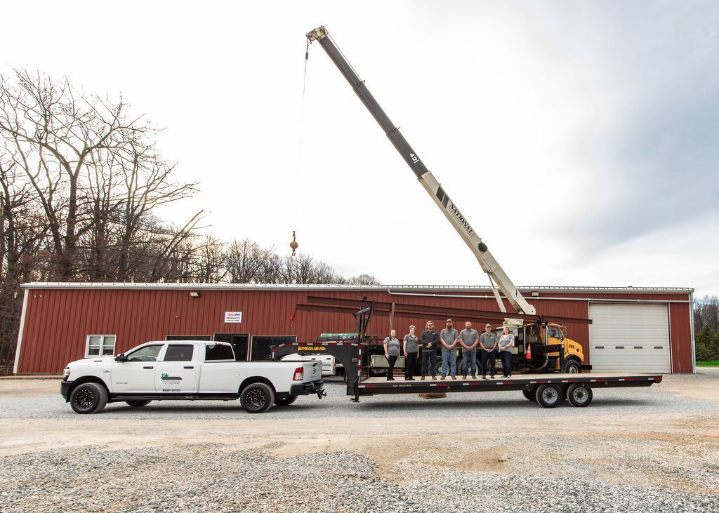 A group of people standing on a flatbed trailer attached to a white pickup truck, with a boom crane parked behind them.
