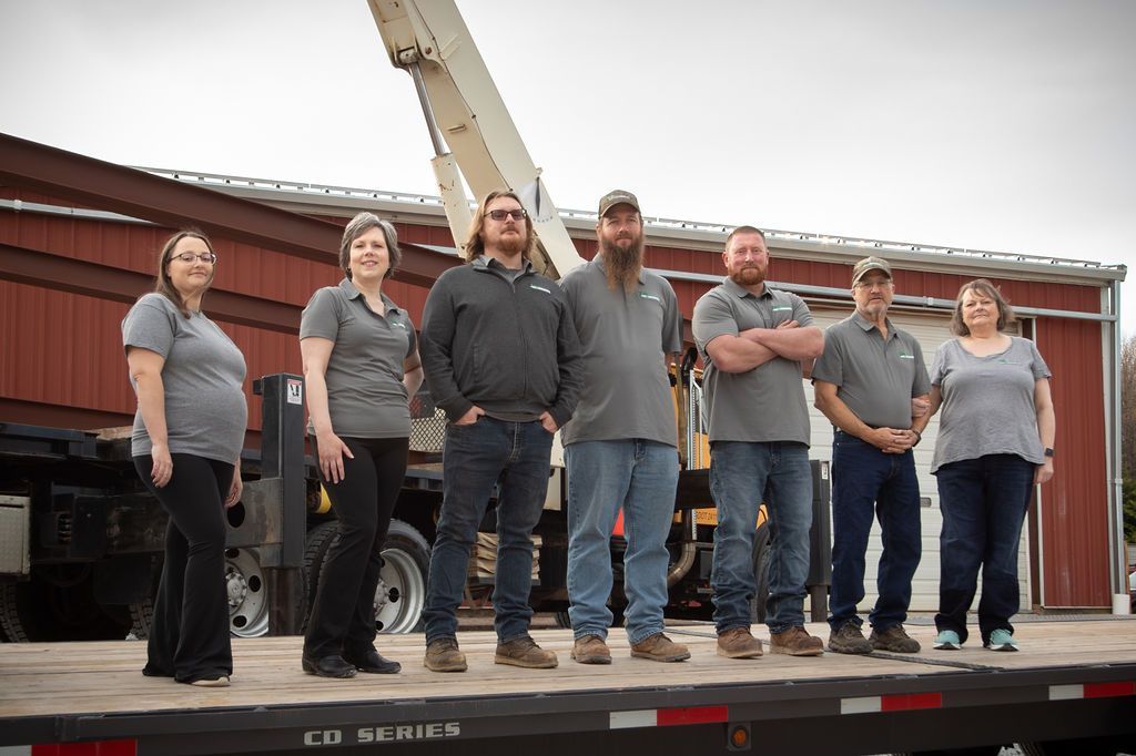 Seven people in grey polo shirts and jeans stand in a row on a trailer bed in front of a red building and crane truck.