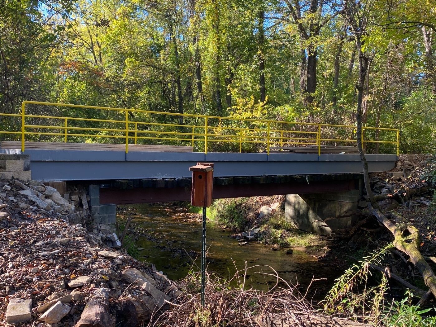 A small, gray wooden bridge with yellow railings spans a shallow creek, with a birdhouse mounted on a pole in the foreground.