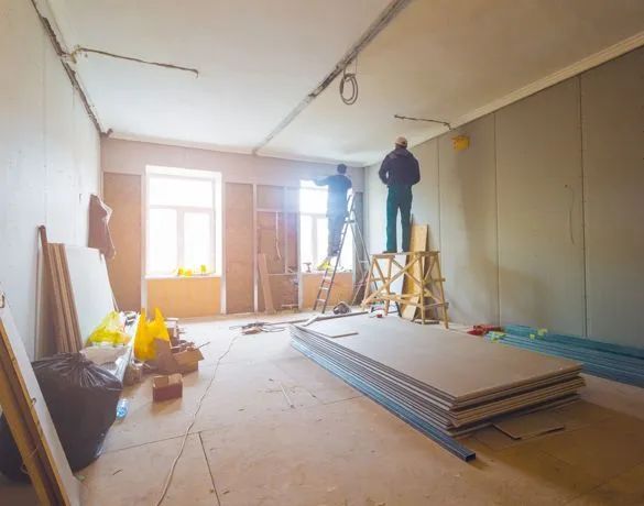 Renovation in progress in a bright room with workers, ladders, tools, and stacked drywall sheets.