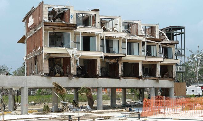 Partially demolished multi-story building with exposed concrete floors and rubble in front, behind orange barriers