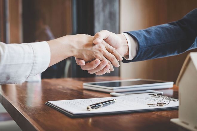 Two people shaking hands across a desk with a pen, notebook, and tablet in a meeting room