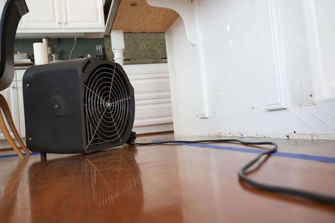 Black portable fan on a wooden floor beside a white wall with a cord trailing across the room