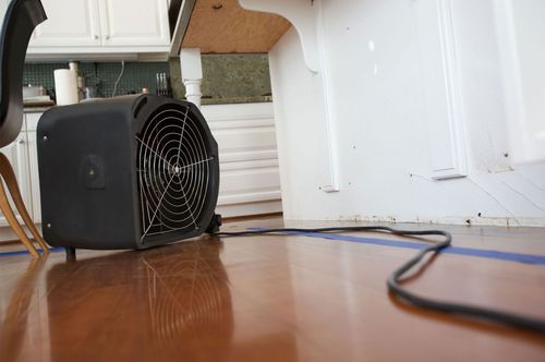 Black portable fan on a wooden floor beside a white wall with a cord trailing across the room