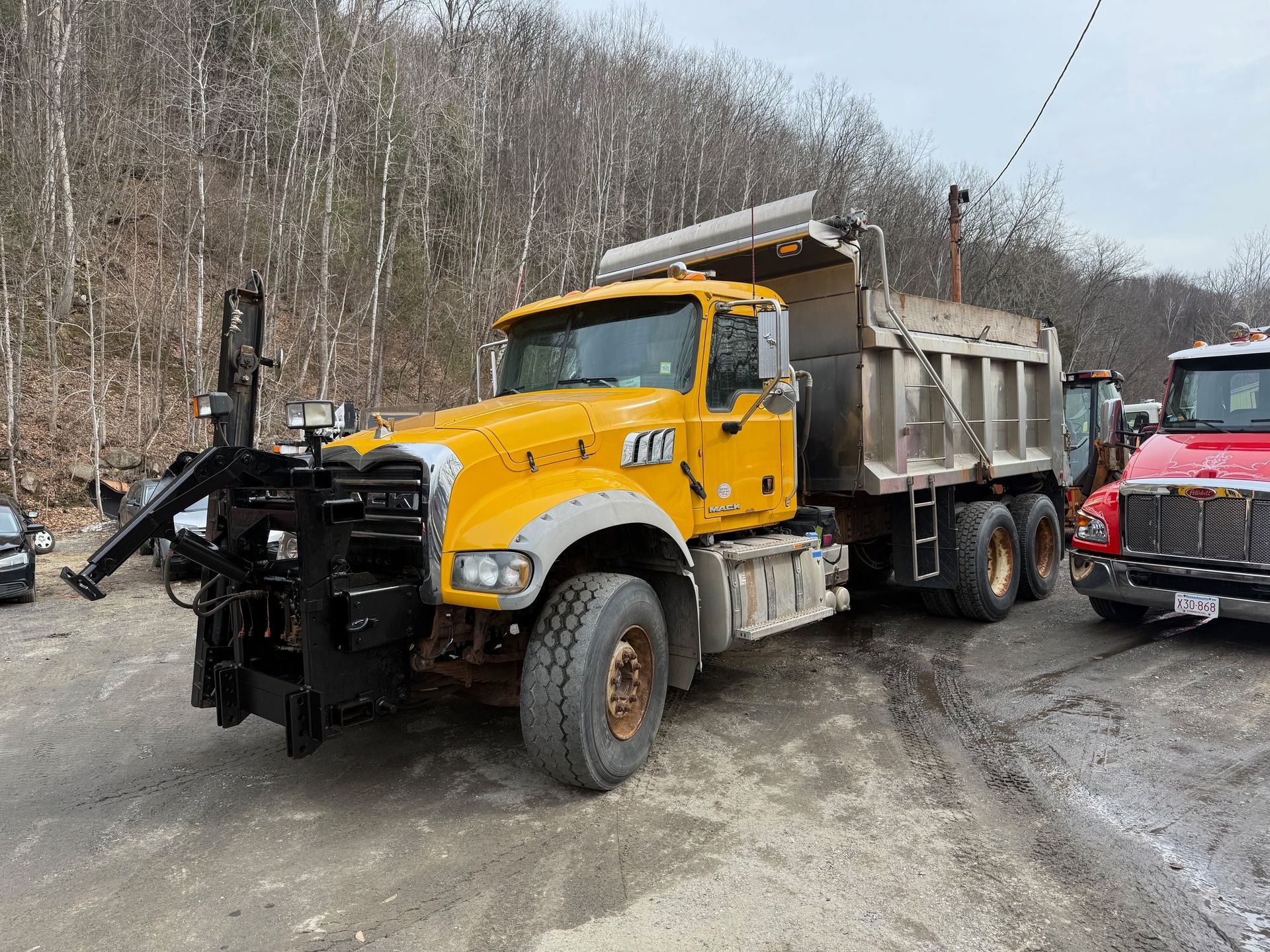 A yellow dump truck with a snow plow mount attached to its front, parked in a lot beside a red truck.