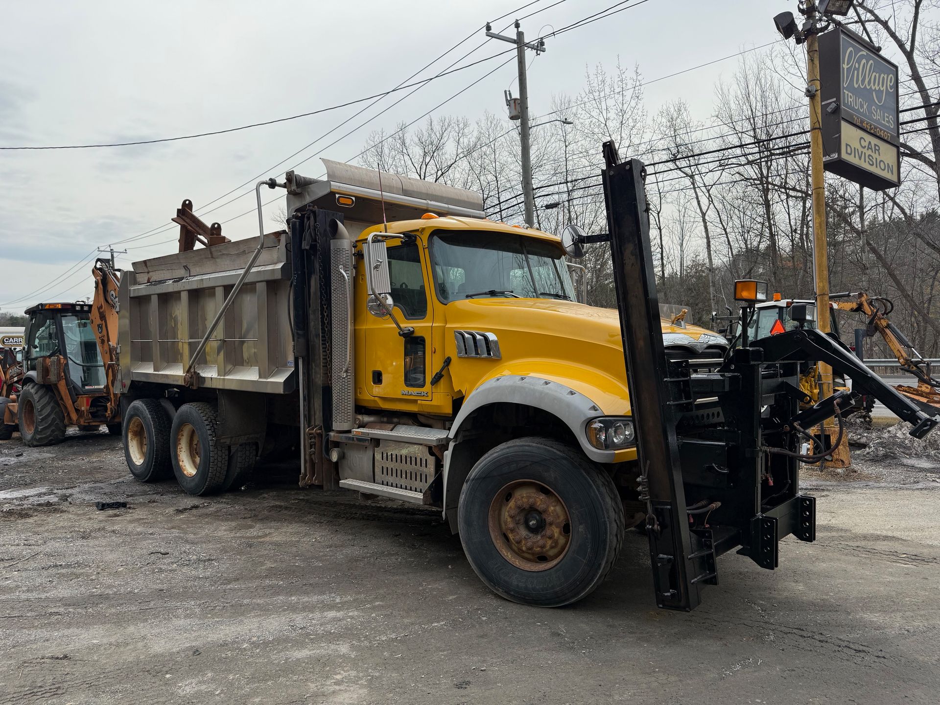A bright yellow dump truck with a front-mounted snowplow hitch parked on a gravel lot under an overcast sky.