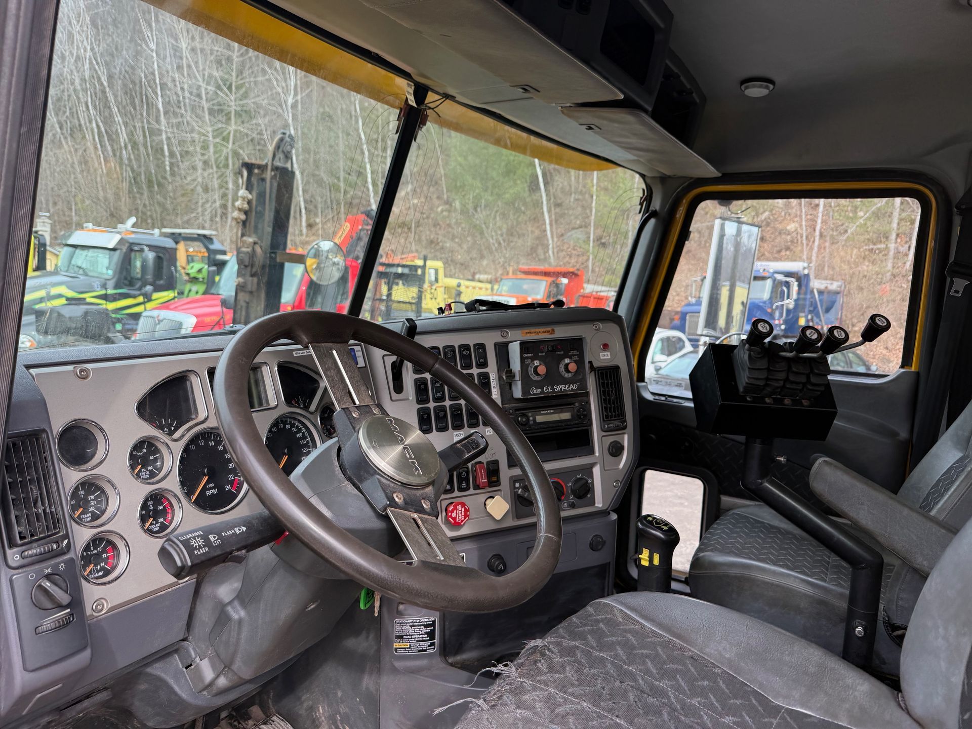 Interior view of a truck cab, showing the steering wheel, instrument panel, and passenger seat with woods in the background.