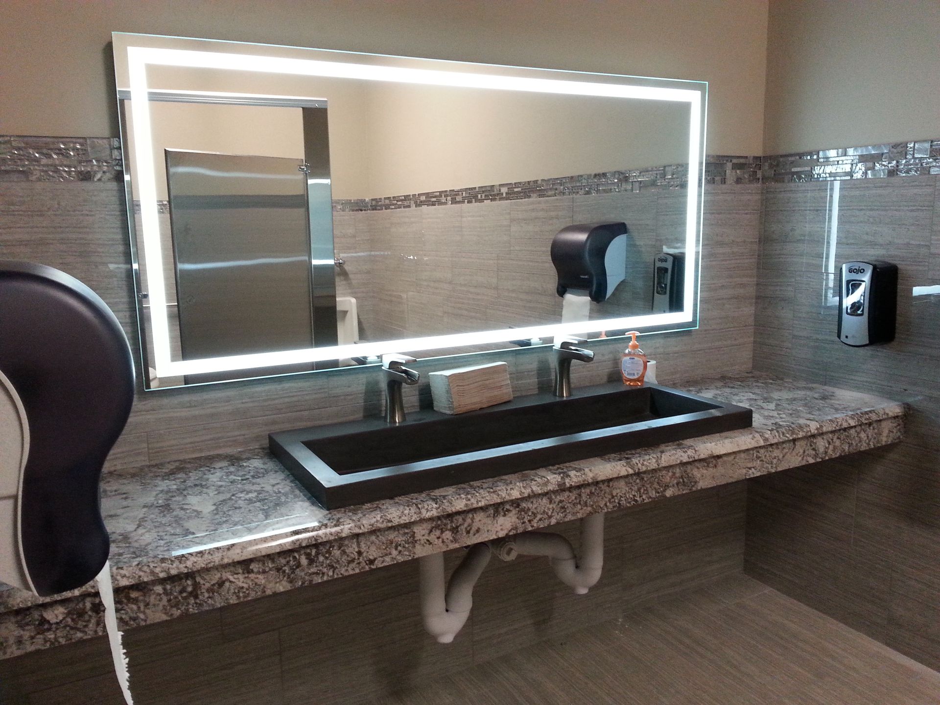 Bathroom sink with a rectangular black basin, large lighted mirror, soap dispenser, and paper towel dispenser.