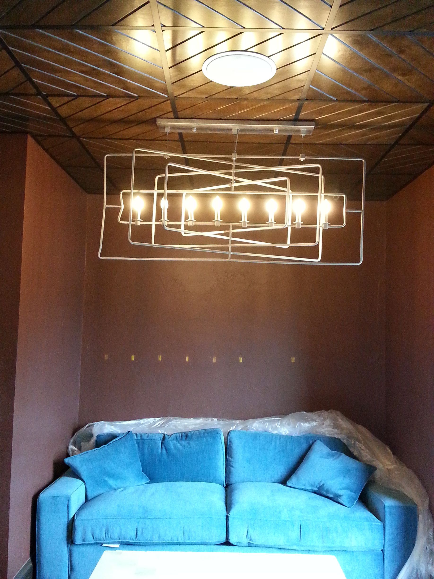 Blue sofa under a geometric light fixture, in a room with a brown wall and wood-patterned ceiling.
