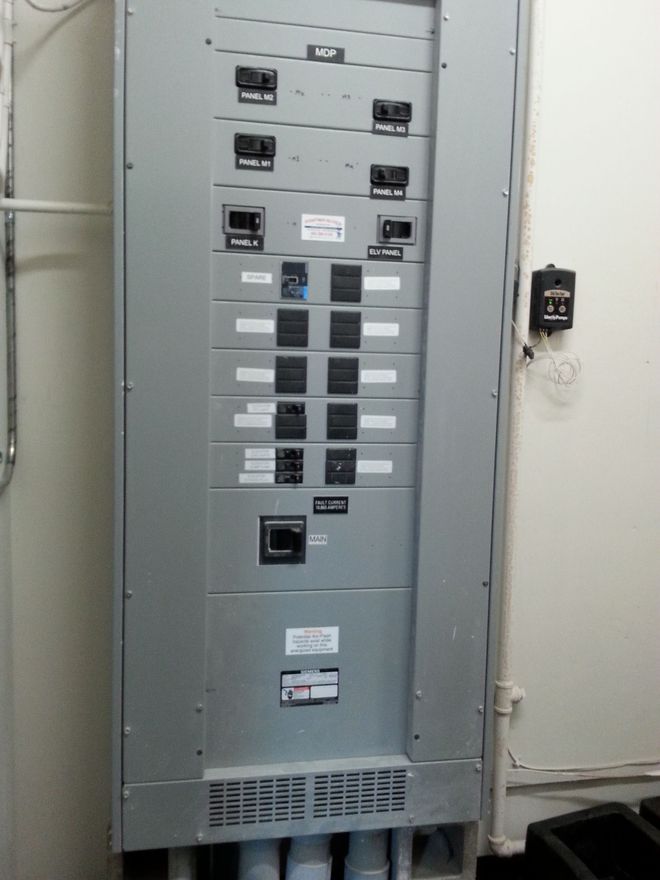 Gray electrical panel with multiple circuit breakers and conduit pipes.