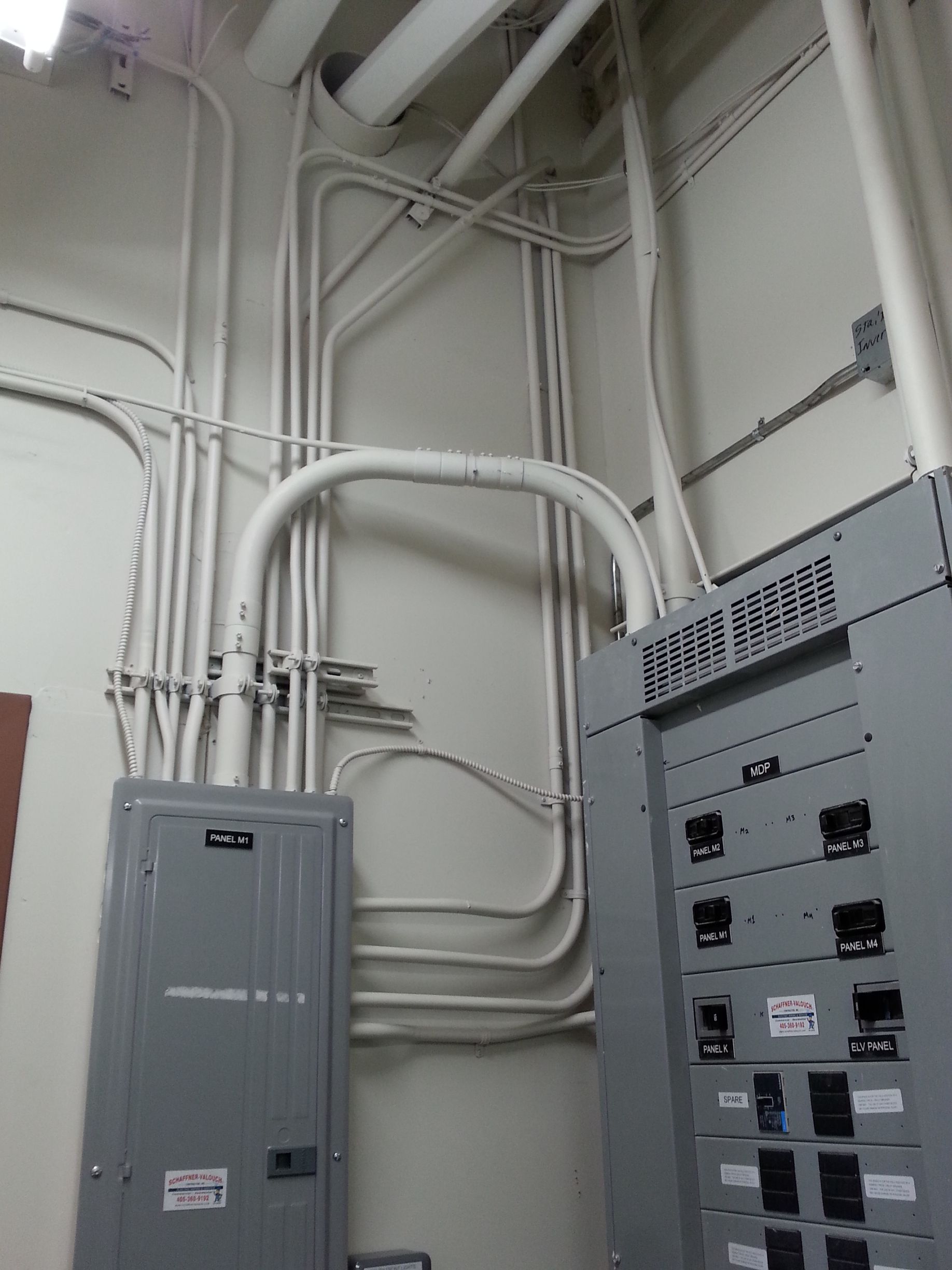 Electrical conduits on a wall with breaker boxes; white pipes, grey boxes, fluorescent lighting.