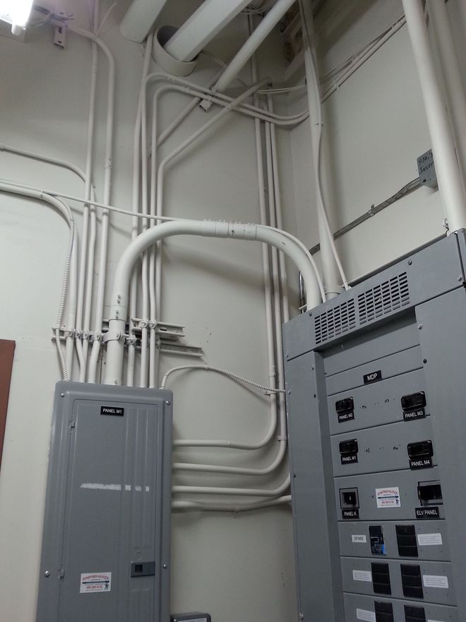Electrical conduits on a wall with breaker boxes; white pipes, grey boxes, fluorescent lighting.