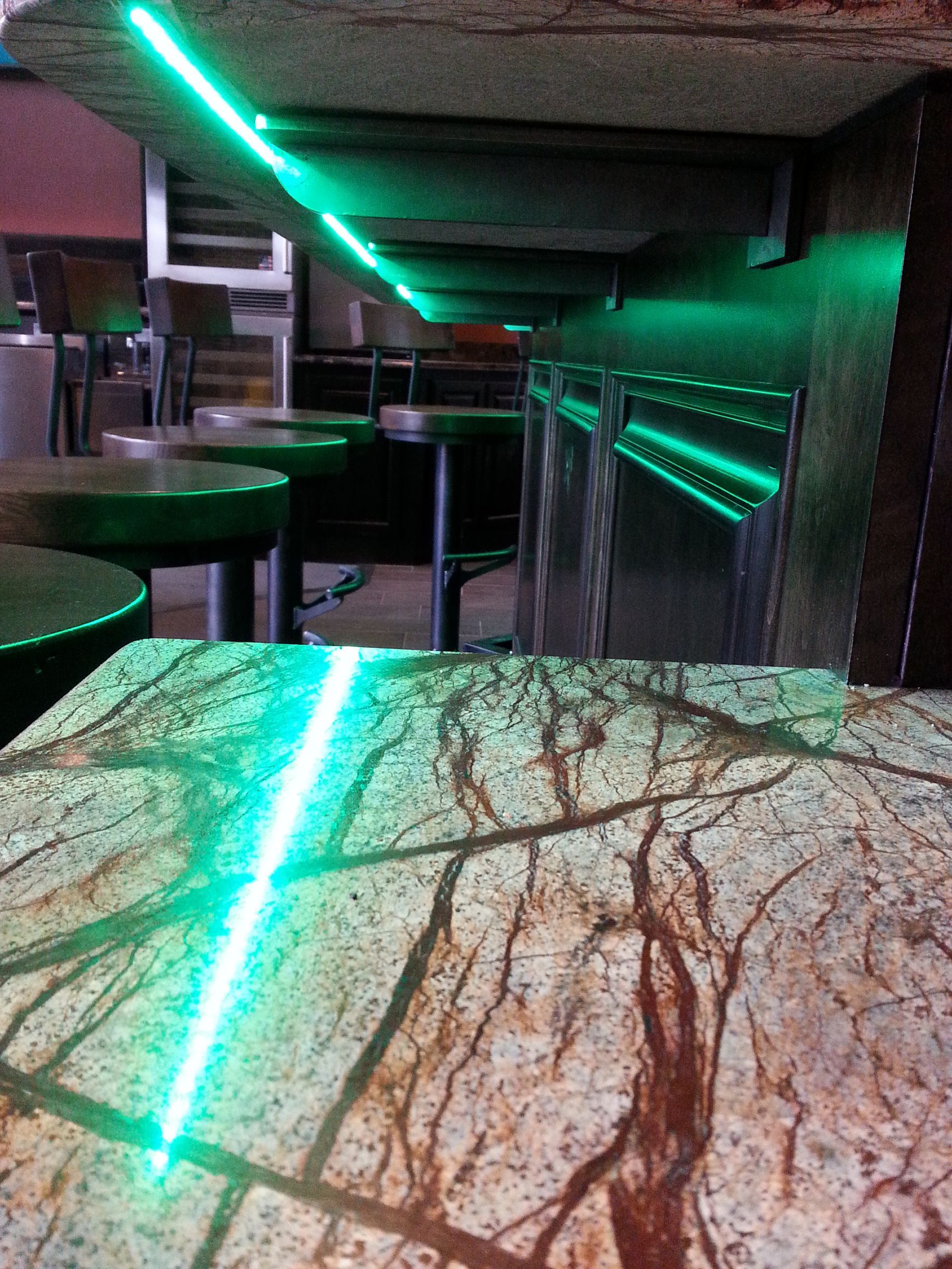 Bar interior with green neon lighting. Tables, stools, and metal surfaces are visible, reflecting the lights.