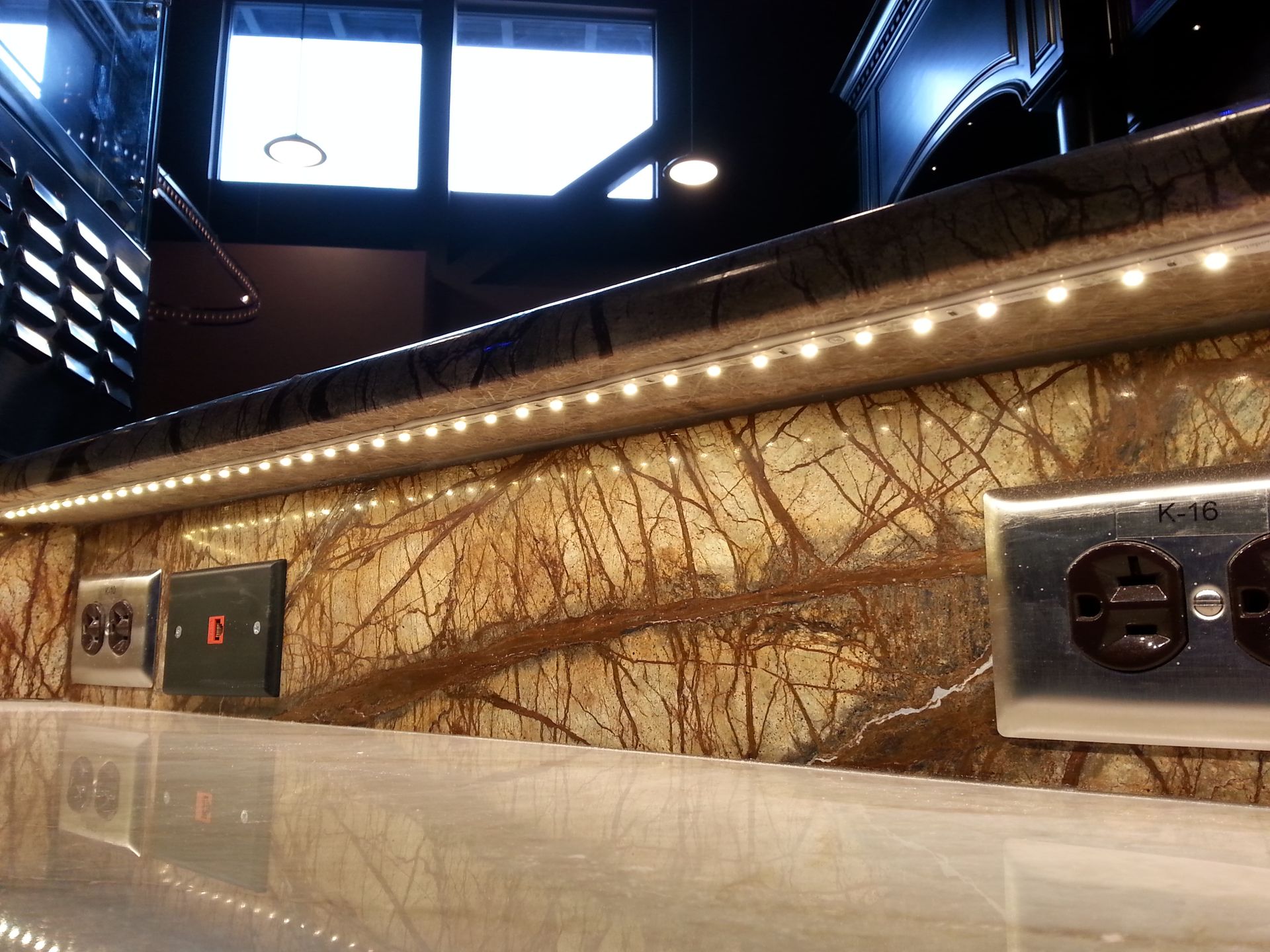 Close-up of a counter with embedded LED lights illuminating a brown and beige patterned backsplash and outlets.