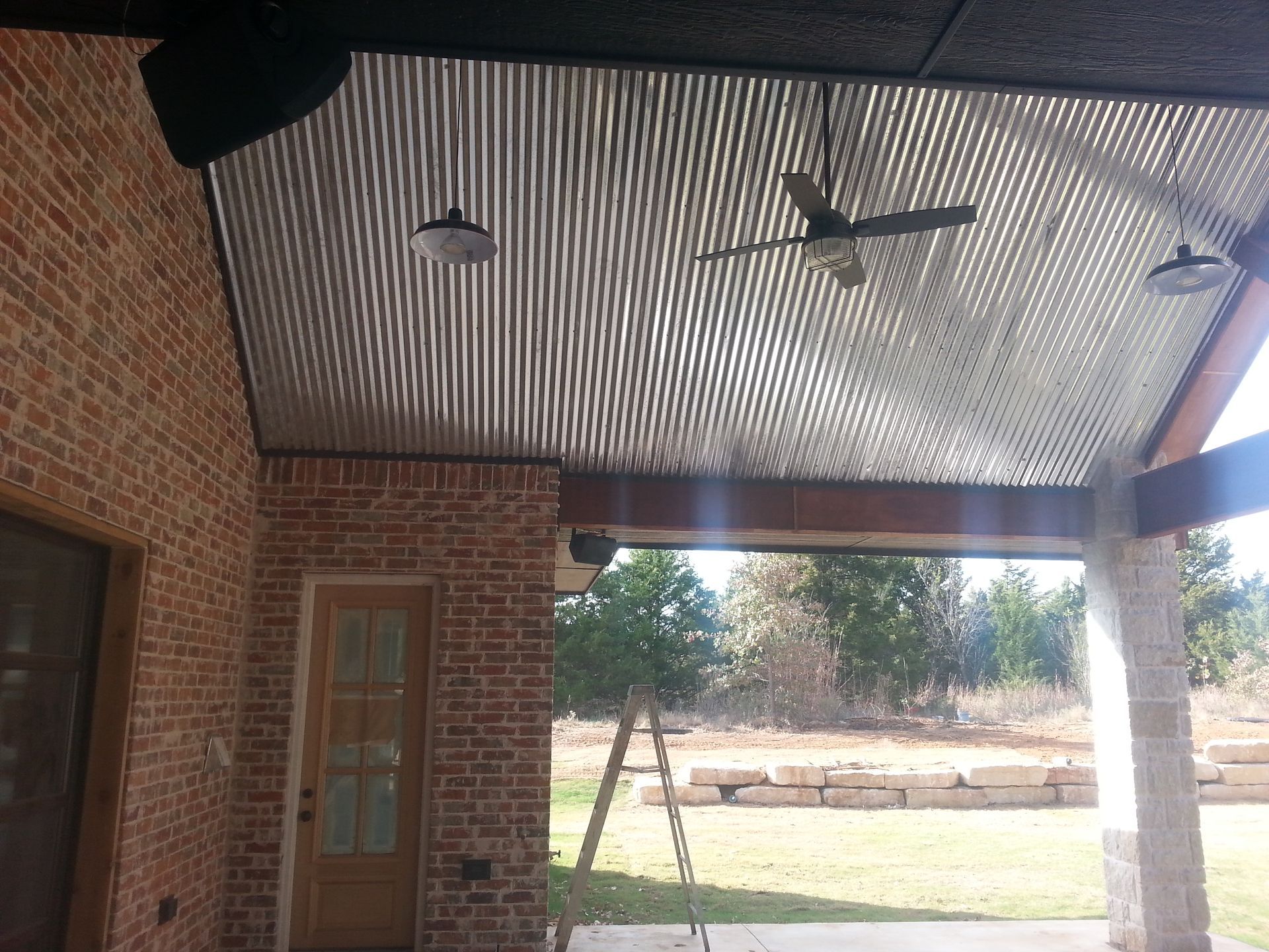 Covered patio with corrugated metal ceiling, brick walls, ceiling fan, and outdoor view.