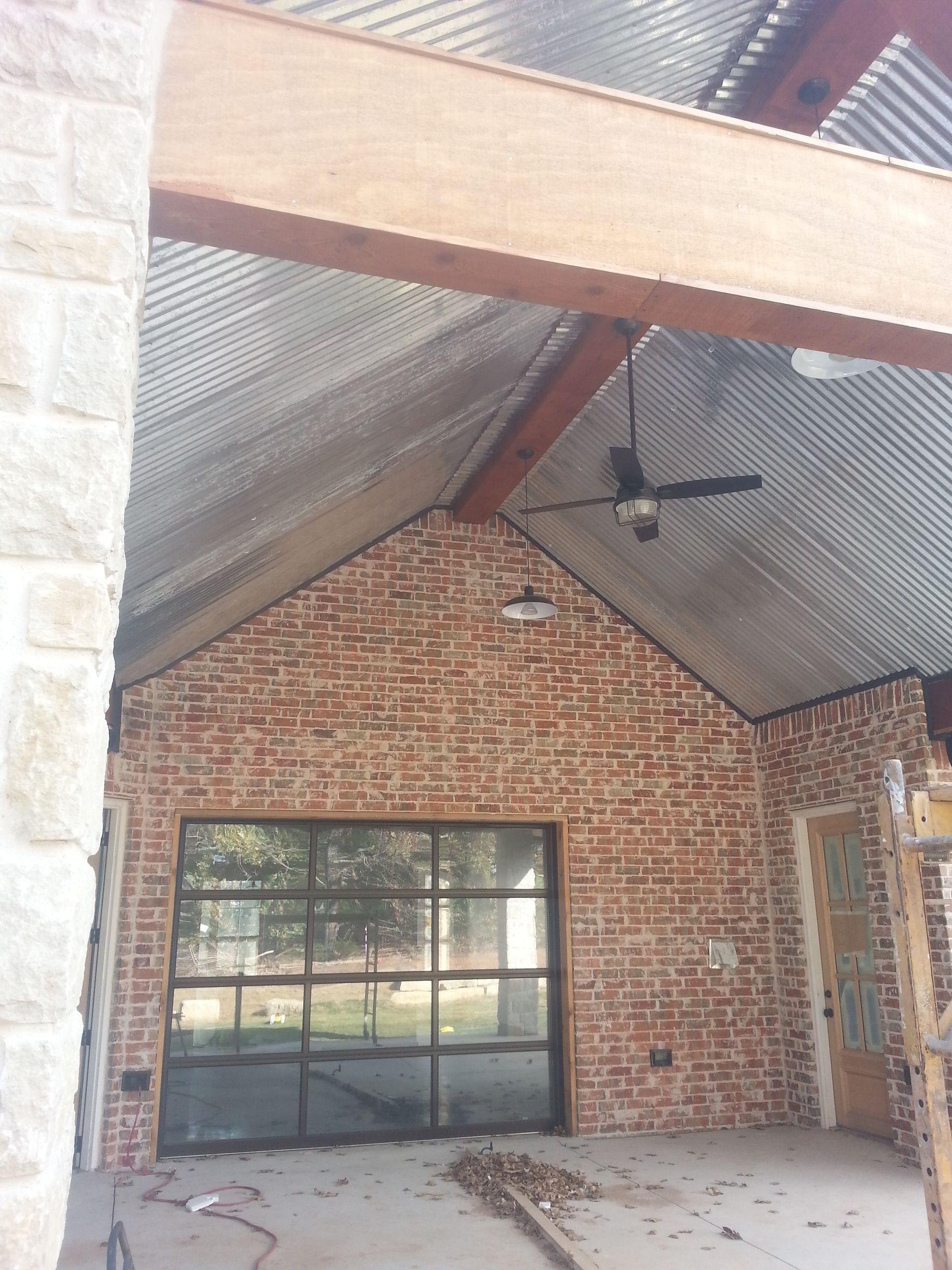 Brick-walled outdoor space with corrugated metal roofing, black-framed glass garage door, and wooden beams.