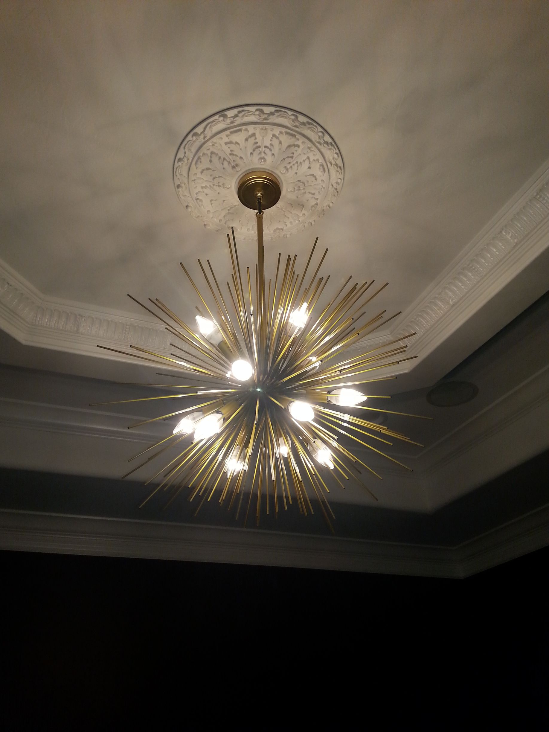 Gold starburst chandelier hanging from a white ceiling with ornate trim.