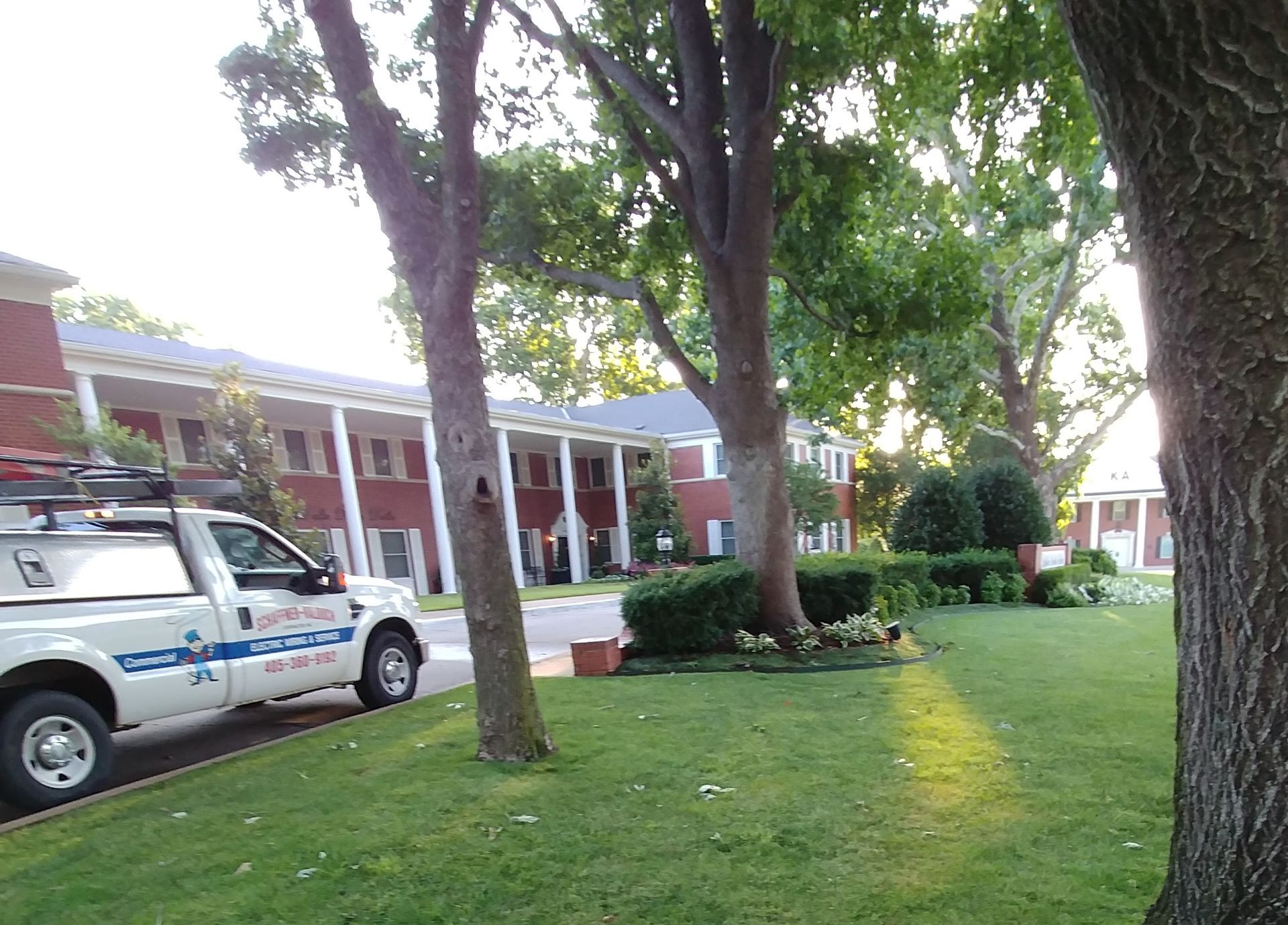 White work truck parked on a grassy lawn in front of a brick building with white columns and large trees.