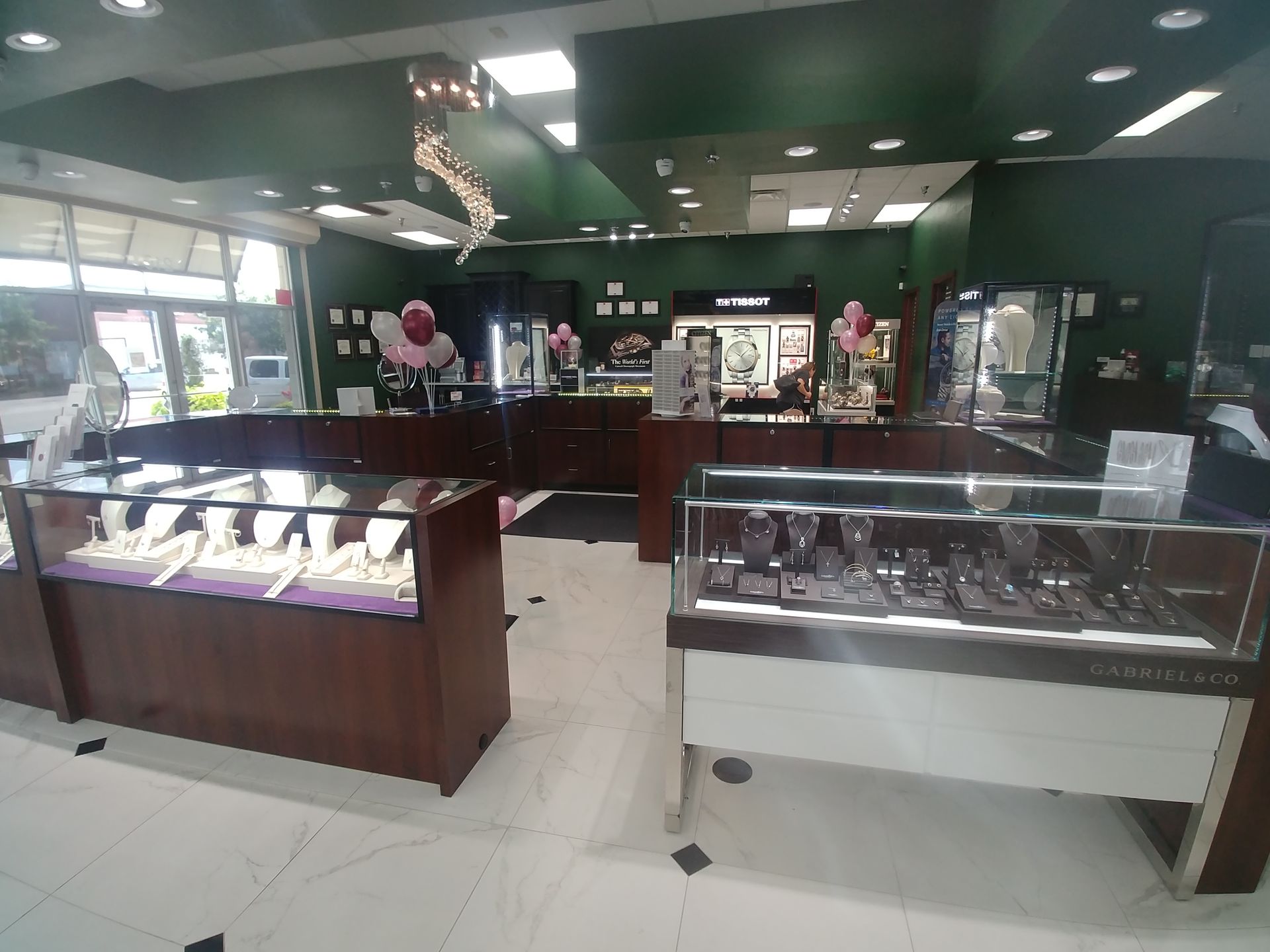 Jewelry store interior with display cases, dark wood, and green walls. Overhead lighting, merchandise visible.