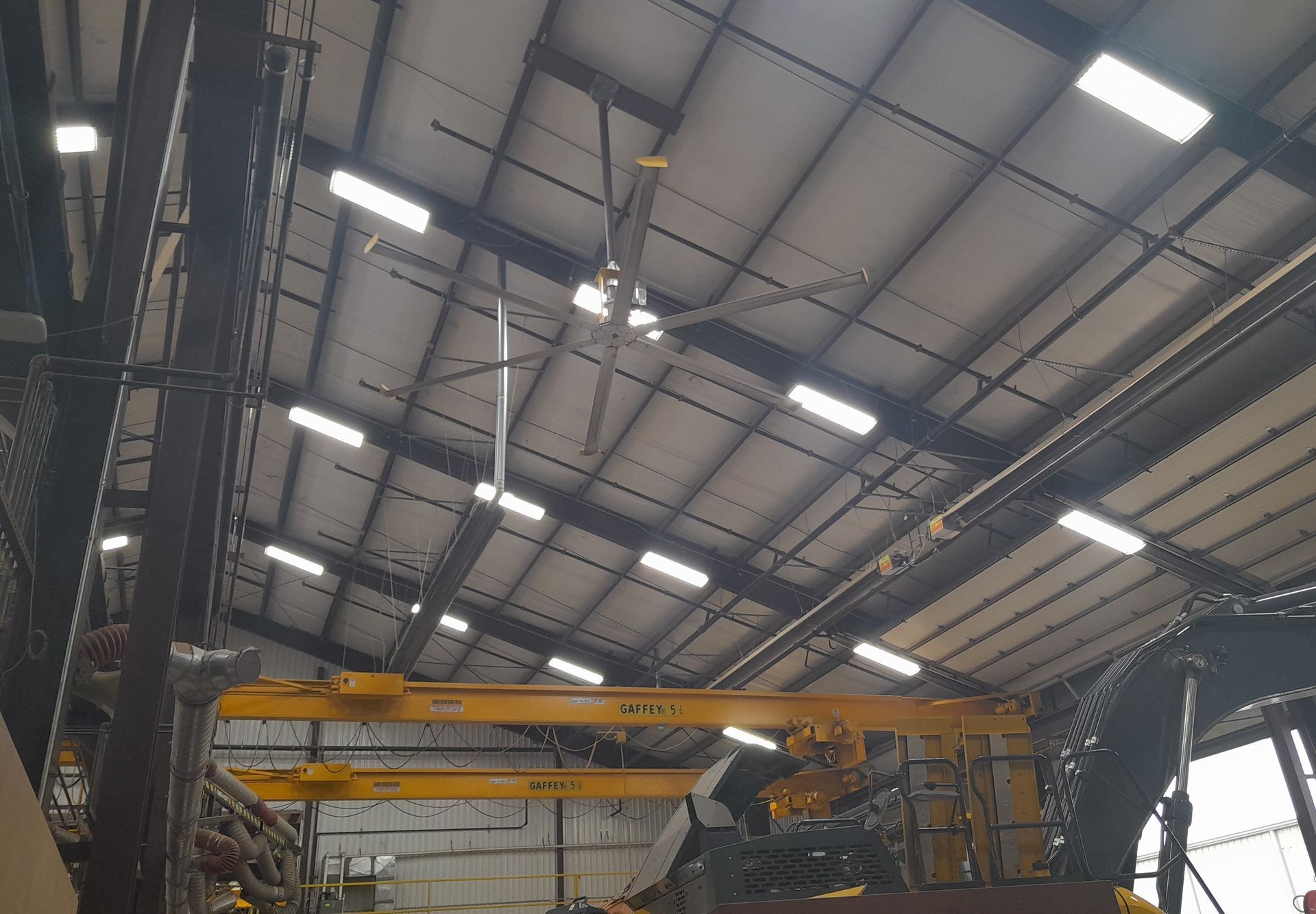 Large industrial fan in a warehouse setting, with bright lights on the ceiling and a yellow crane below.