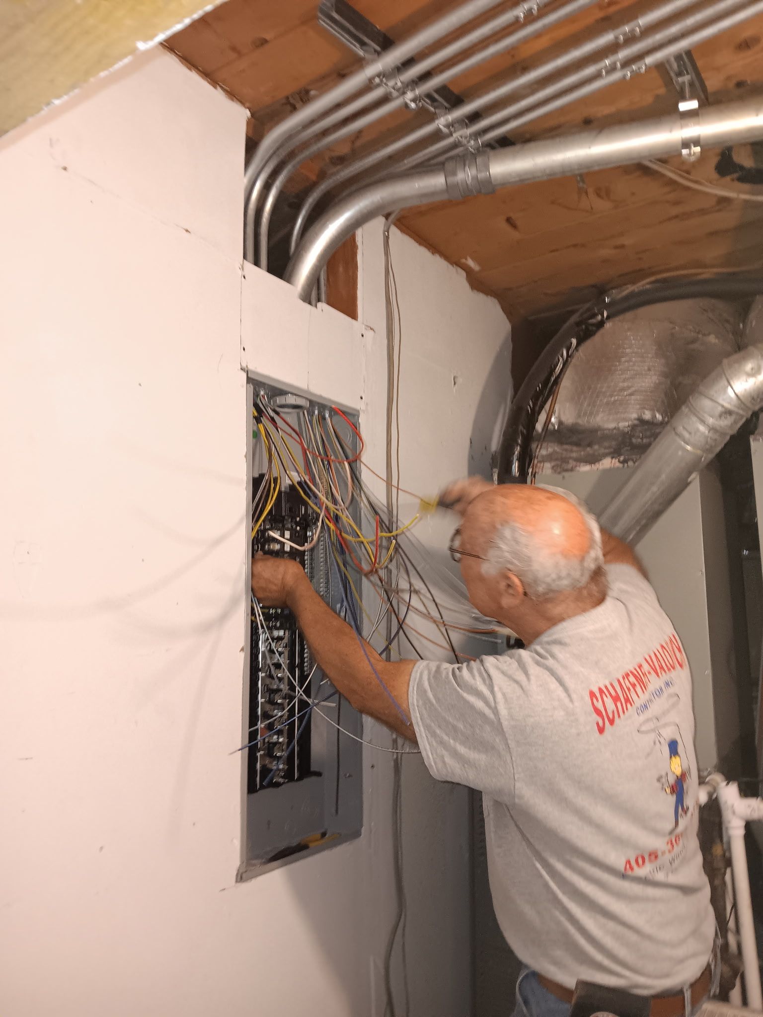 Man working on electrical panel, wiring visible. Conduit overhead, drywall surrounds.