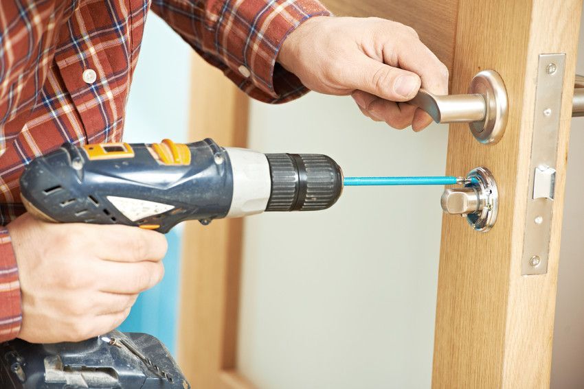 Person using a power drill to install a silver doorknob on a light wood door.