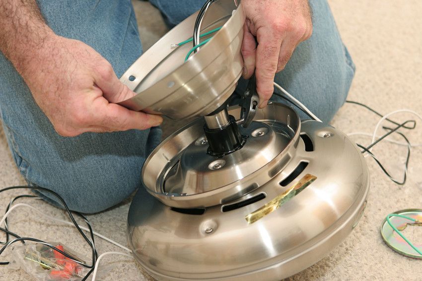 Person in jeans assembling a brushed nickel ceiling fan, connecting wires. Indoors, close-up shot.