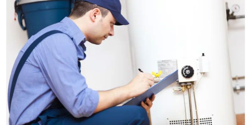 A plumber in a blue uniform and cap checks a water heater, writing on a clipboard.