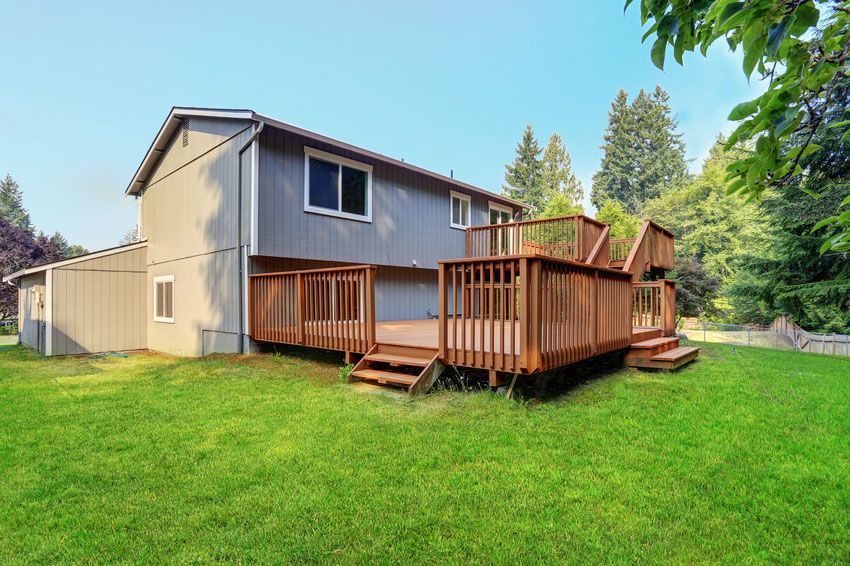 Two-story gray house with a large wooden deck, surrounded by green grass and trees under a blue sky.