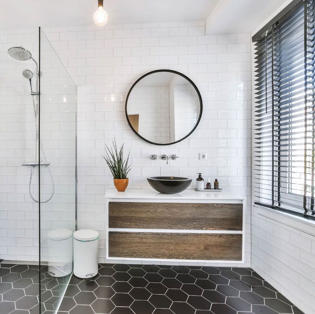 Modern bathroom with black hexagonal tile floor, white brick-like walls, a wooden vanity, and a round mirror.