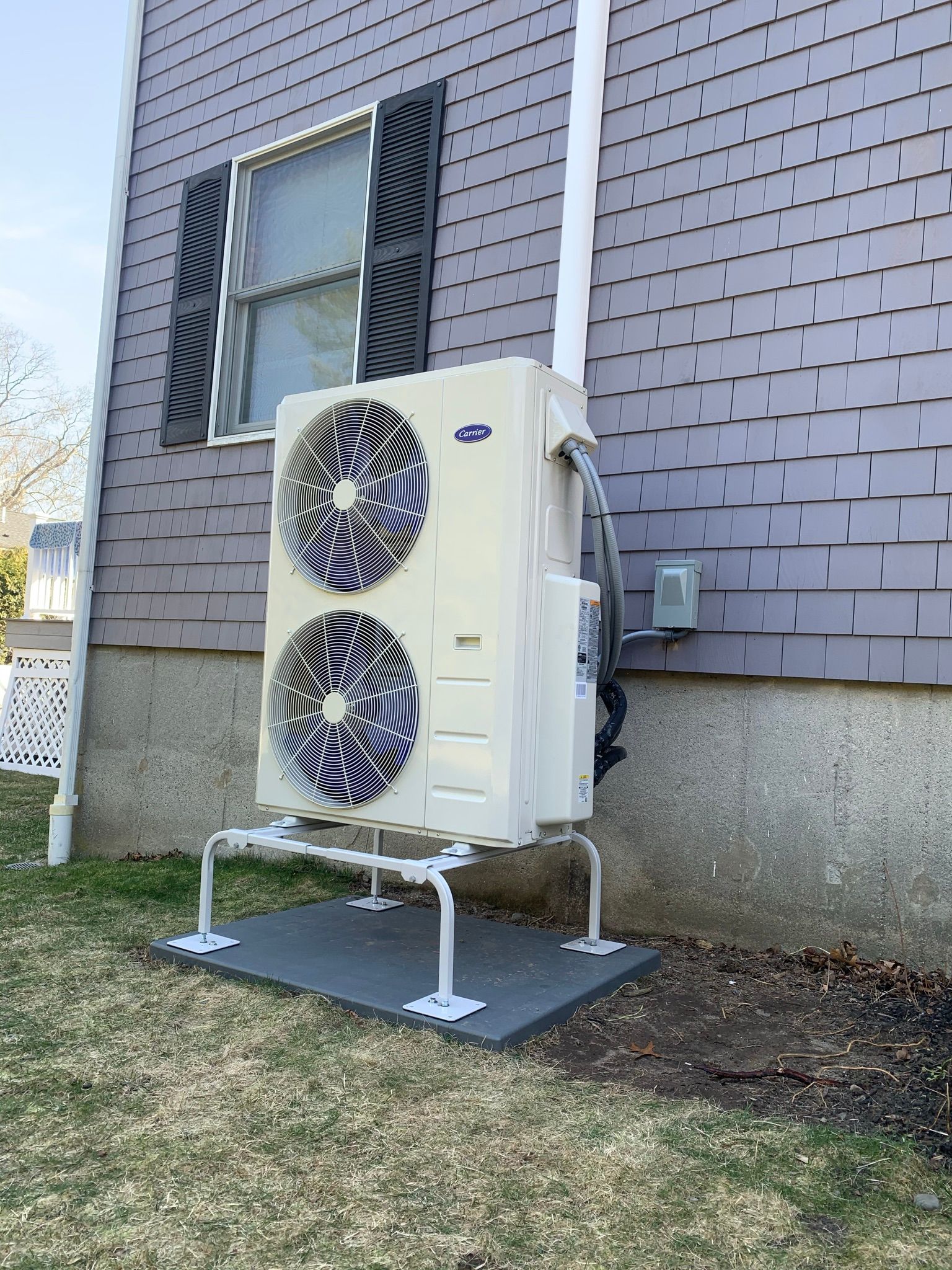 Air conditioner unit mounted on legs next to a house with gray siding.