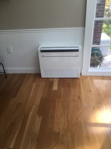 White air conditioner unit on wood floor, near a window and wall with white wainscoting.