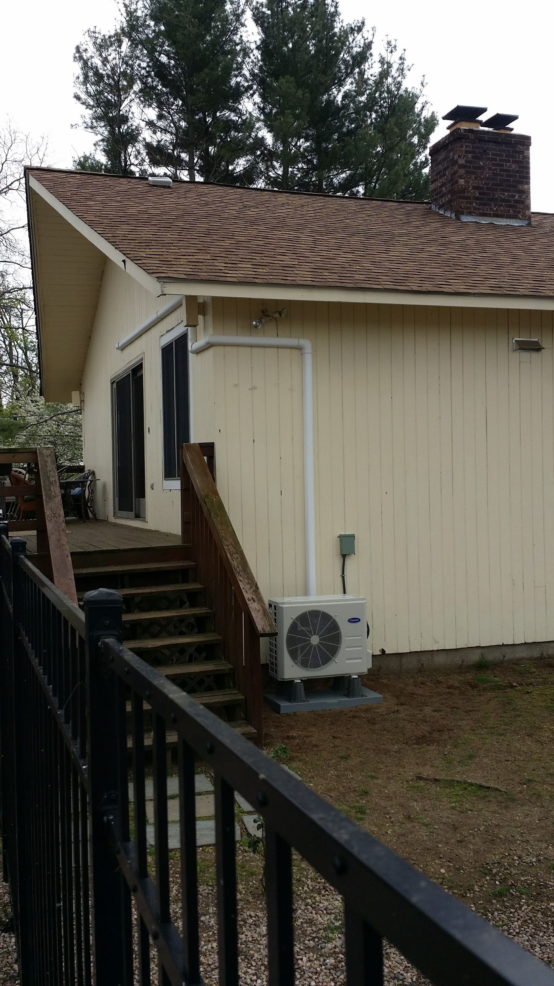 A yellow house with brown roof and a wooden deck, black railing, and outdoor AC unit.